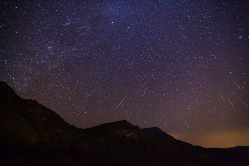 A starry night sky filled with numerous shooting stars over dark mountain silhouettes.