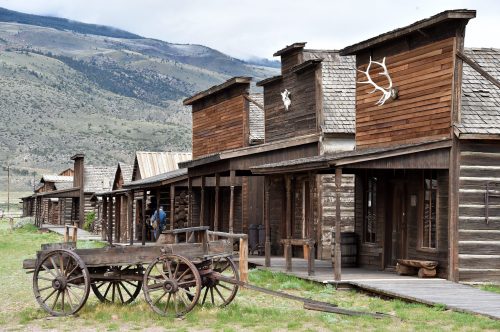 A rustic old western town with wooden buildings and a wagon in front, set against a mountainous backdrop.