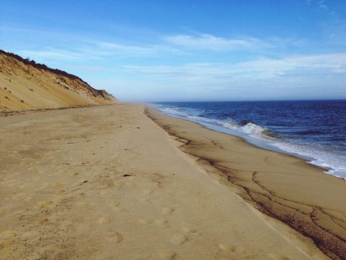A serene beach scene with soft sand, gentle waves, and a clear blue sky, bordered by a sandy cliff.