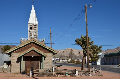 A small, rustic church with a steeple, surrounded by desert landscape and power lines under a clear blue sky.