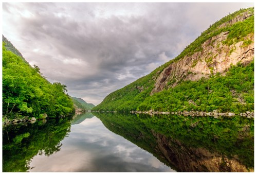 A serene lake surrounded by lush green mountains under a cloudy sky, reflecting the landscape in calm waters.