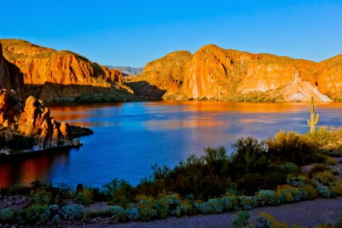 A serene lake surrounded by red rock formations and desert vegetation under a clear blue sky at sunset.