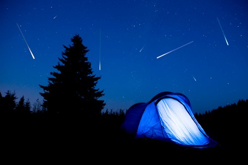 A glowing tent under a starry night sky with multiple shooting stars and a silhouette of a tree.