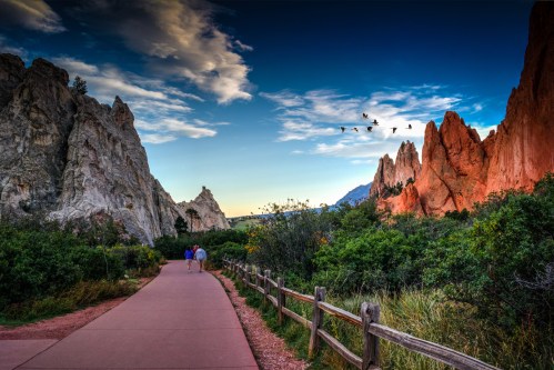 A scenic pathway through rocky terrain, with two people walking and birds flying in a colorful sky.