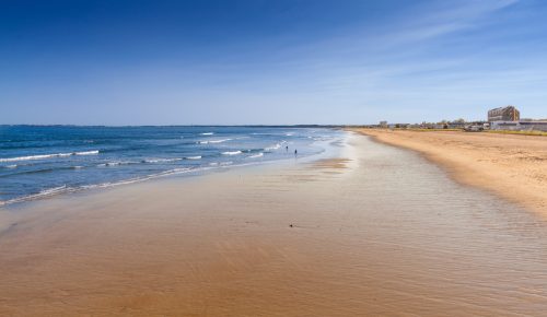 A serene beach scene with gentle waves, golden sand, and a clear blue sky, featuring a distant shoreline and a few people.