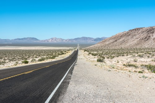 A long, straight road stretches through a desert landscape, flanked by mountains under a clear blue sky.