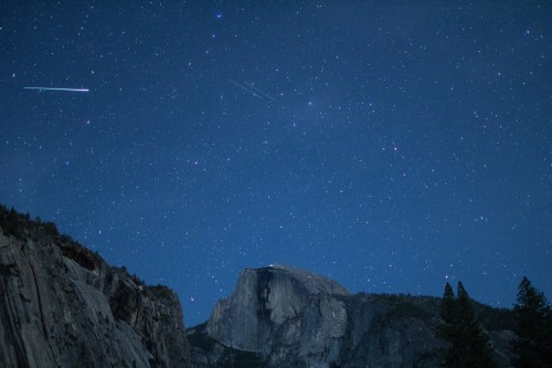 A starry night sky over a mountain, with bright stars and a few streaks of light from passing objects.