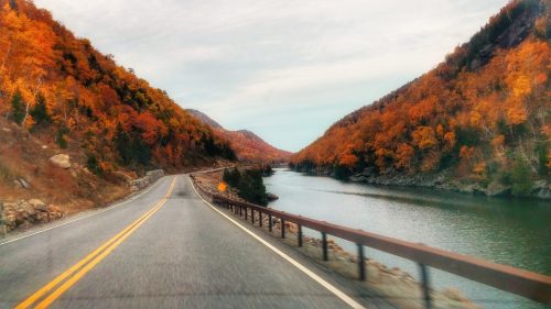 A scenic road winds alongside a river, framed by vibrant autumn foliage on both sides.