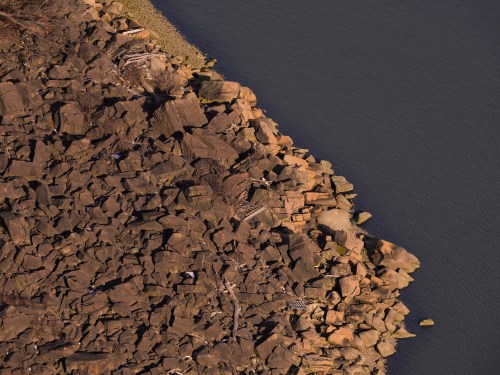 Aerial view of a rocky shoreline meeting calm water, with scattered debris along the edge.