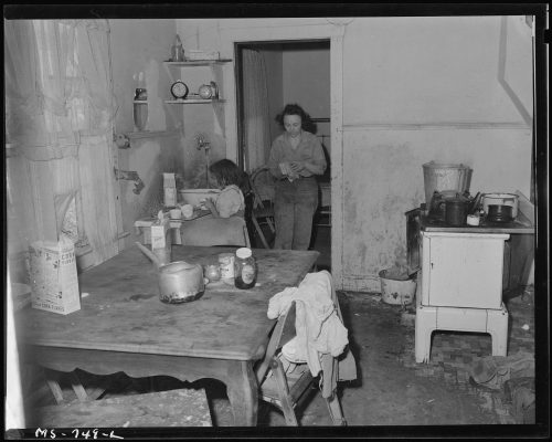 A dimly lit kitchen with two women, one seated at a table and the other standing, surrounded by everyday household items.