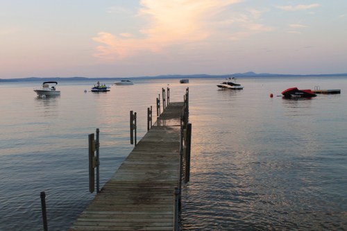 A wooden dock extends into calm water, with several boats anchored nearby under a pastel sunset sky.