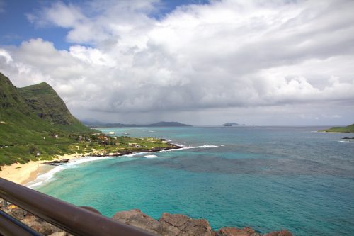 A scenic view of a coastline with green mountains, turquoise water, and a cloudy sky in the background.