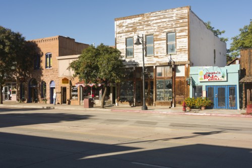 A sunny street scene featuring rustic buildings, including a grill, with trees lining the sidewalk.