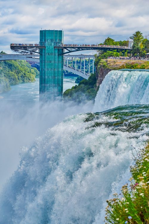 A view of Niagara Falls with a observation deck above, surrounded by mist and lush greenery.