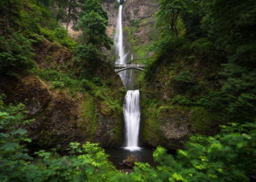 A stunning waterfall cascades down rocky cliffs, surrounded by lush greenery and a bridge above.
