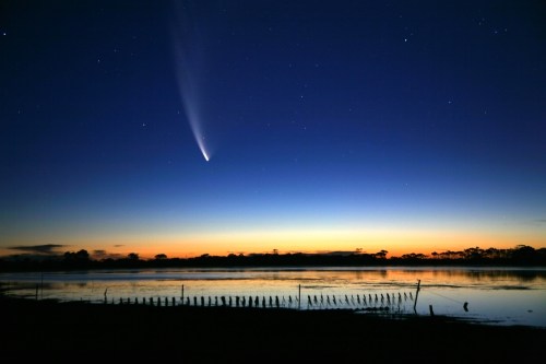 A comet streaks across a twilight sky above a calm lake, reflecting the colors of sunset and stars.