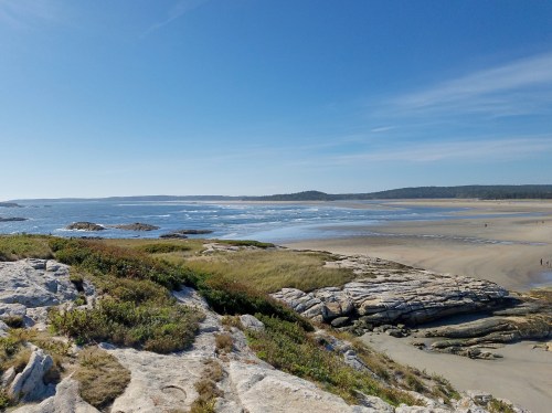 A scenic beach view with rocky formations, gentle waves, and a clear blue sky in the background.