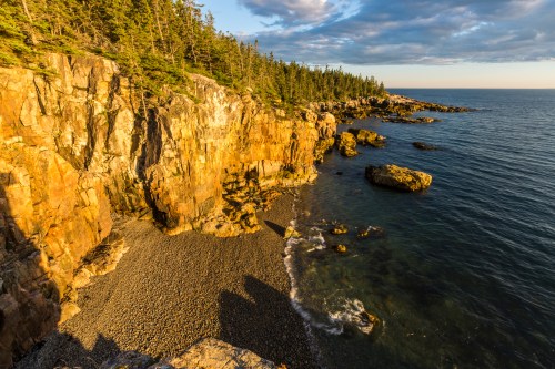 A rocky coastline with cliffs and trees, overlooking a calm sea under a partly cloudy sky at sunset.