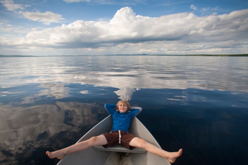 A boy relaxes in a small boat on a calm lake, surrounded by clouds reflecting on the water's surface.