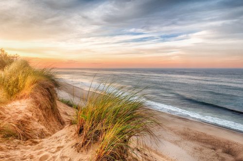 A serene beach scene at sunset, with sandy dunes and tall grass overlooking gentle waves and a colorful sky.