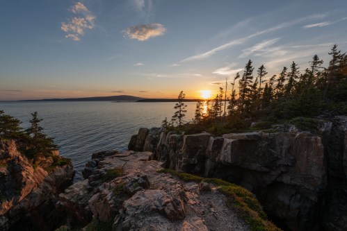 Sunset over a calm sea, with rocky cliffs and trees framing the view, creating a serene coastal landscape.