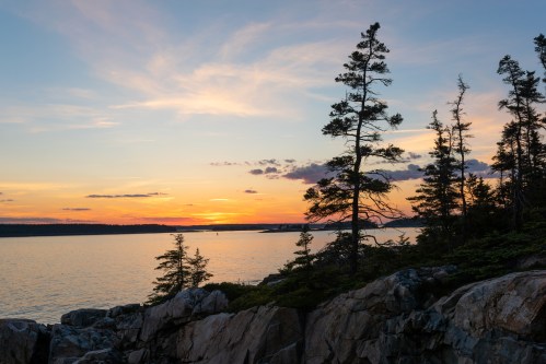 A serene sunset over a calm lake, framed by tall trees and rocky shoreline, with colorful clouds in the sky.