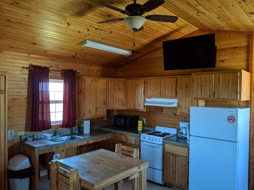 Cozy wooden kitchen with a ceiling fan, table, appliances, and a TV mounted on the wall. Natural light from a window.