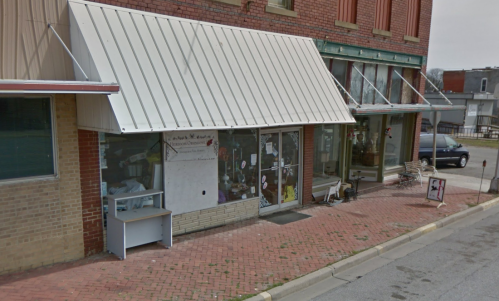 A storefront with a white awning, large windows, and a brick facade, featuring a sidewalk and nearby parked car.