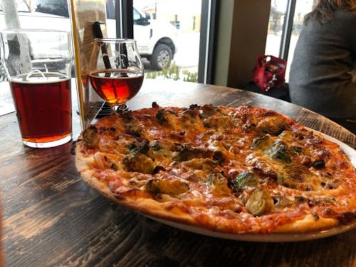 A pizza topped with vegetables sits on a wooden table next to two glasses of beer, with a snowy scene outside.