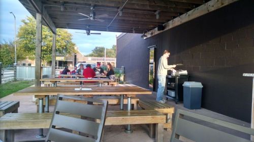 Outdoor dining area with wooden tables, a person grilling, and a group of people enjoying a meal in the background.