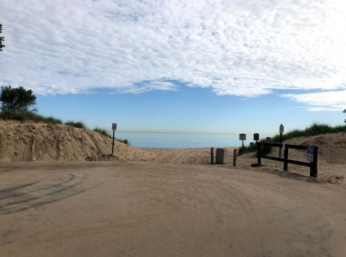 A sandy path leads to a calm beach under a cloudy sky, with signs and a wooden fence on either side.