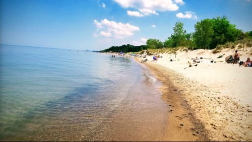 A serene beach scene with calm water, sandy shore, and people relaxing under a clear blue sky with scattered clouds.