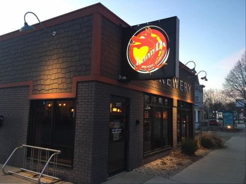 Exterior of a brewery with a glowing sign, featuring a dragon logo, at dusk.