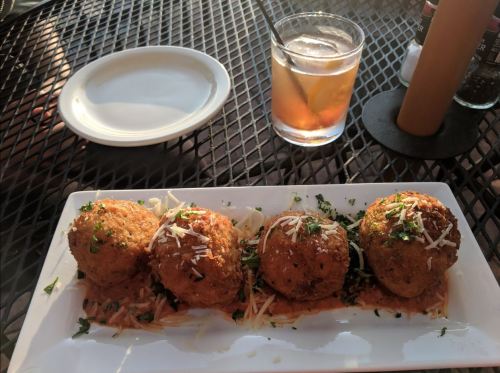 Three golden-brown fried balls on a plate with sauce and garnish, alongside a glass of drink on a table.