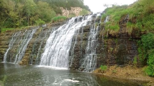 A cascading waterfall flows over rocky cliffs into a serene pool, surrounded by lush greenery and trees.