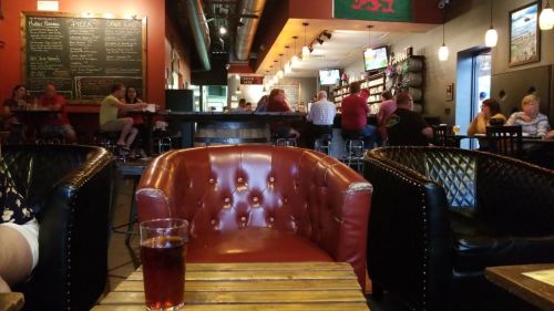 A cozy café interior with leather chairs, wooden tables, and patrons enjoying food and drinks in a lively atmosphere.