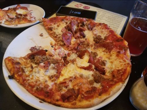 A partially eaten pizza topped with pepperoni, sausage, and cheese on a white plate, with a drink and menu in the background.