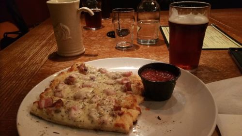 A plate with a slice of pizza topped with cheese and bacon, accompanied by a small cup of marinara sauce and a pint of beer.