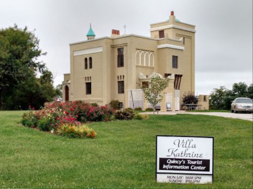 Historic Villa Kathrine, Quincy’s Tourist Information Center, surrounded by greenery and flowers, with a sign in front.