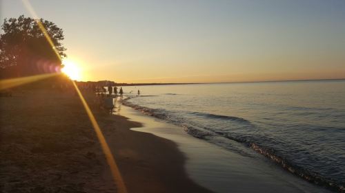 Sunset over a calm beach, with silhouettes of people walking along the shore and gentle waves lapping at the sand.