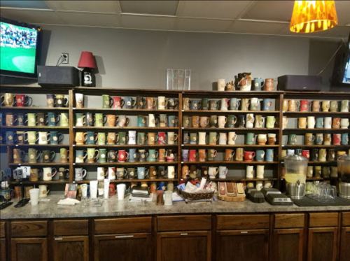A wall of colorful coffee mugs displayed on shelves, with a countertop featuring drinks and snacks. Two TVs are visible.