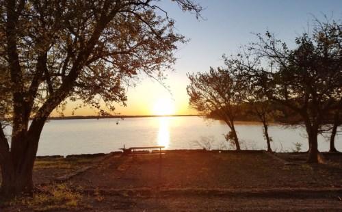 Sunset over a calm lake, framed by trees, with a bench in the foreground and warm golden light reflecting on the water.