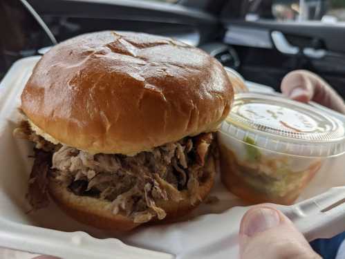 A close-up of a pulled pork sandwich on a bun, held in one hand, with a small container of coleslaw beside it.