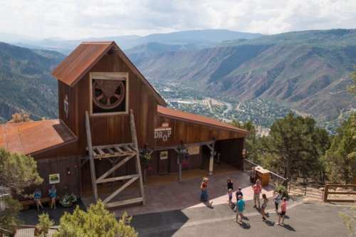 A rustic wooden building on a hillside with a scenic mountain view and visitors exploring the area.