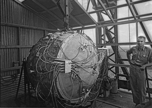 A man stands beside a large, spherical device with numerous wires, inside a metal structure.