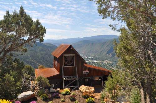 A rustic wooden building on a hillside, surrounded by trees and colorful flowers, overlooking a valley and mountains.