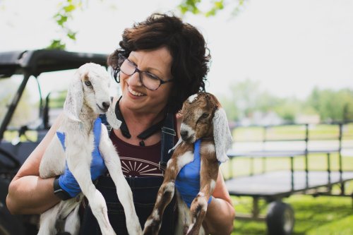 A woman in glasses smiles while holding two baby goats outdoors, wearing gloves and surrounded by greenery.