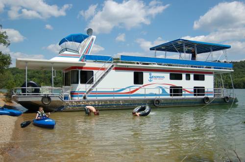 A houseboat docked by the shore with people enjoying the water and kayaks nearby under a blue sky.