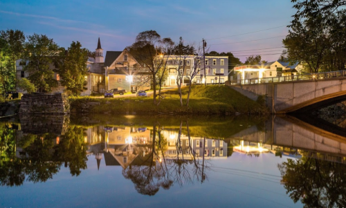 A serene evening scene of a town by a river, with buildings and trees reflecting in the water under a twilight sky.