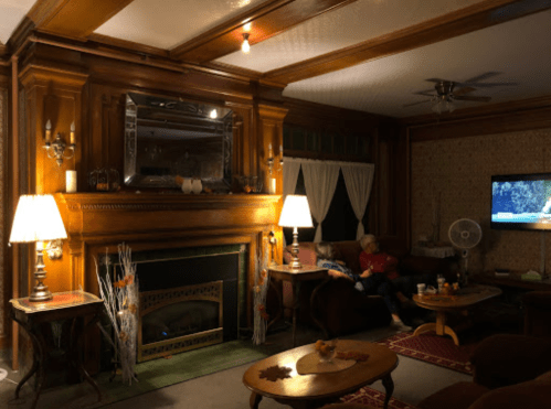 Cozy living room with wooden accents, a fireplace, two people sitting on a couch, and a TV in the background.